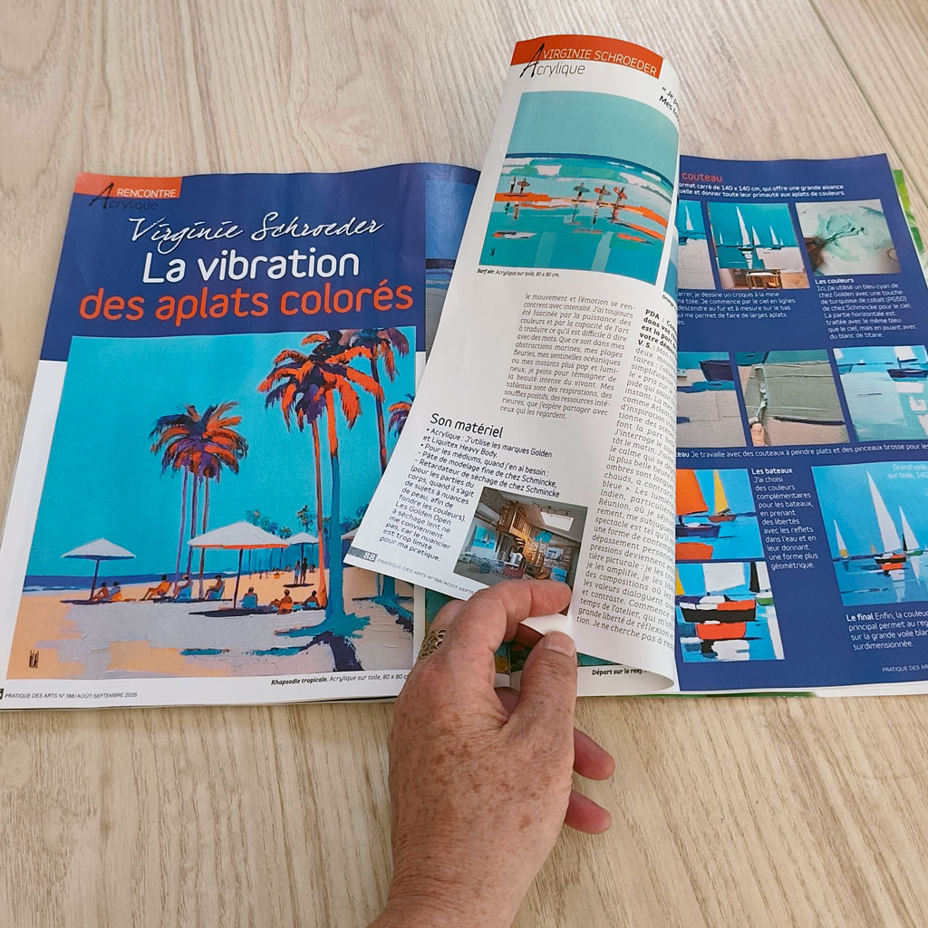 Person turning a page in a colorful art magazine laid on a wooden surface, featuring a tropical beach illustration in blue tones.
